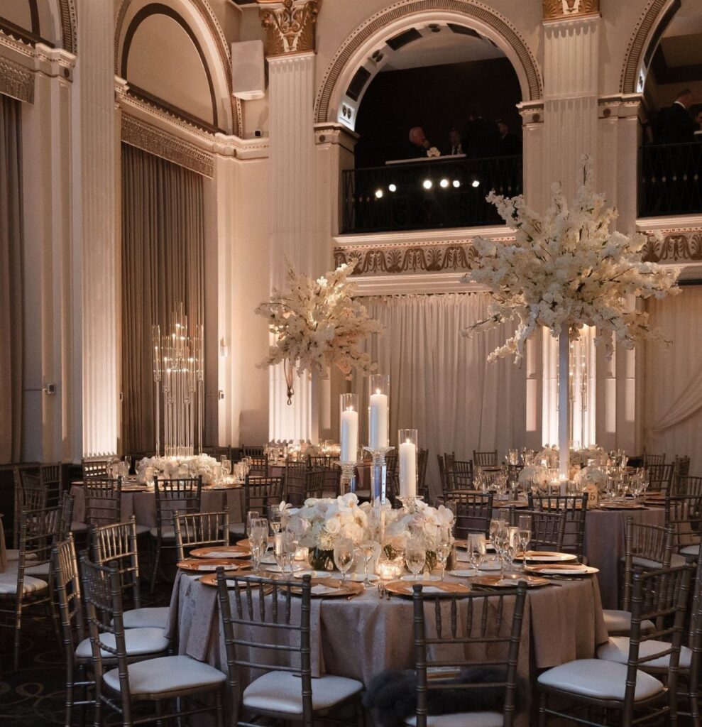 Elegant ballroom wedding reception with round tables, tall white floral centerpieces, and grand architectural columns.