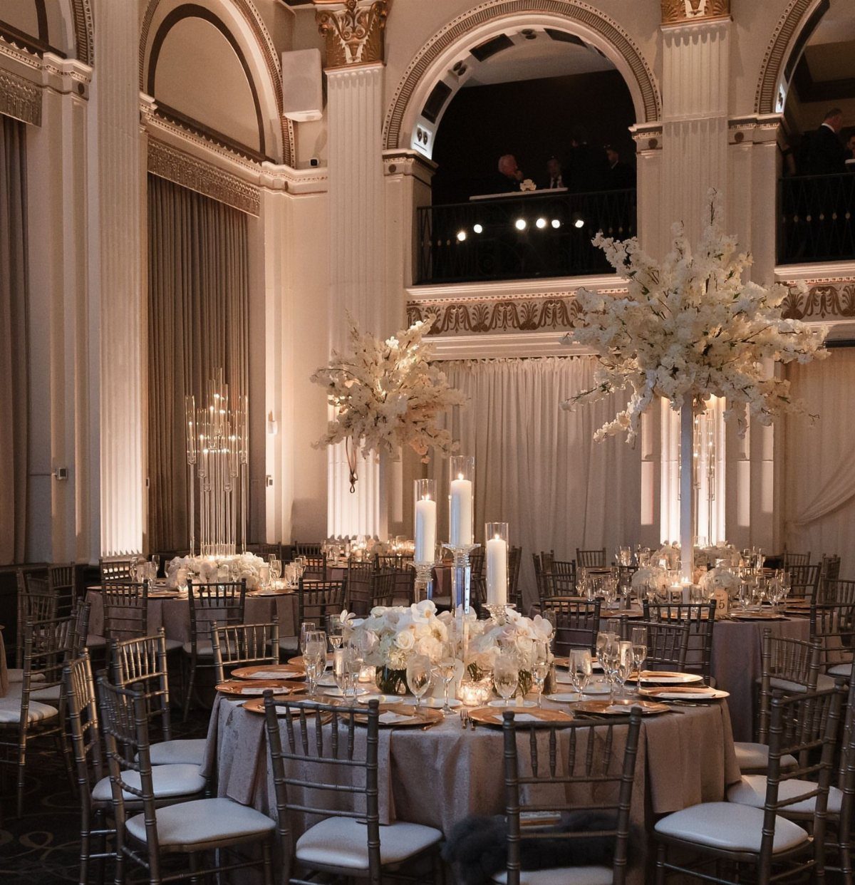 Elegant ballroom wedding reception with round tables, tall white floral centerpieces, and grand architectural columns.