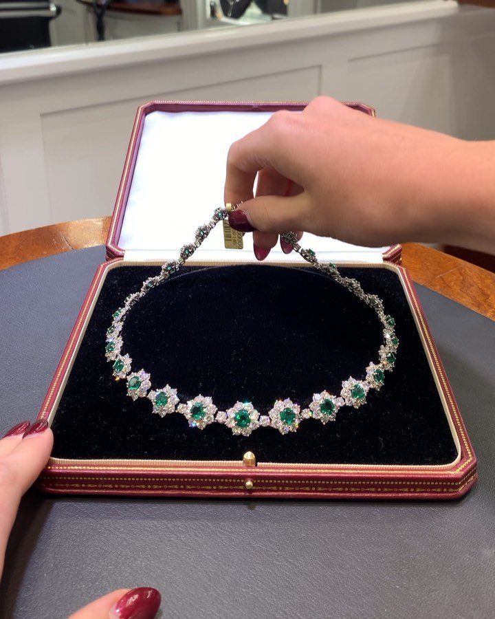 Hands holding an open jewelry box displaying a diamond and emerald necklace.
