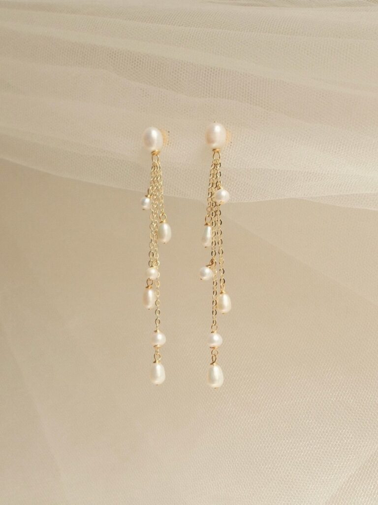 Long gold chain earrings with hanging white pearls against a cream fabric background.