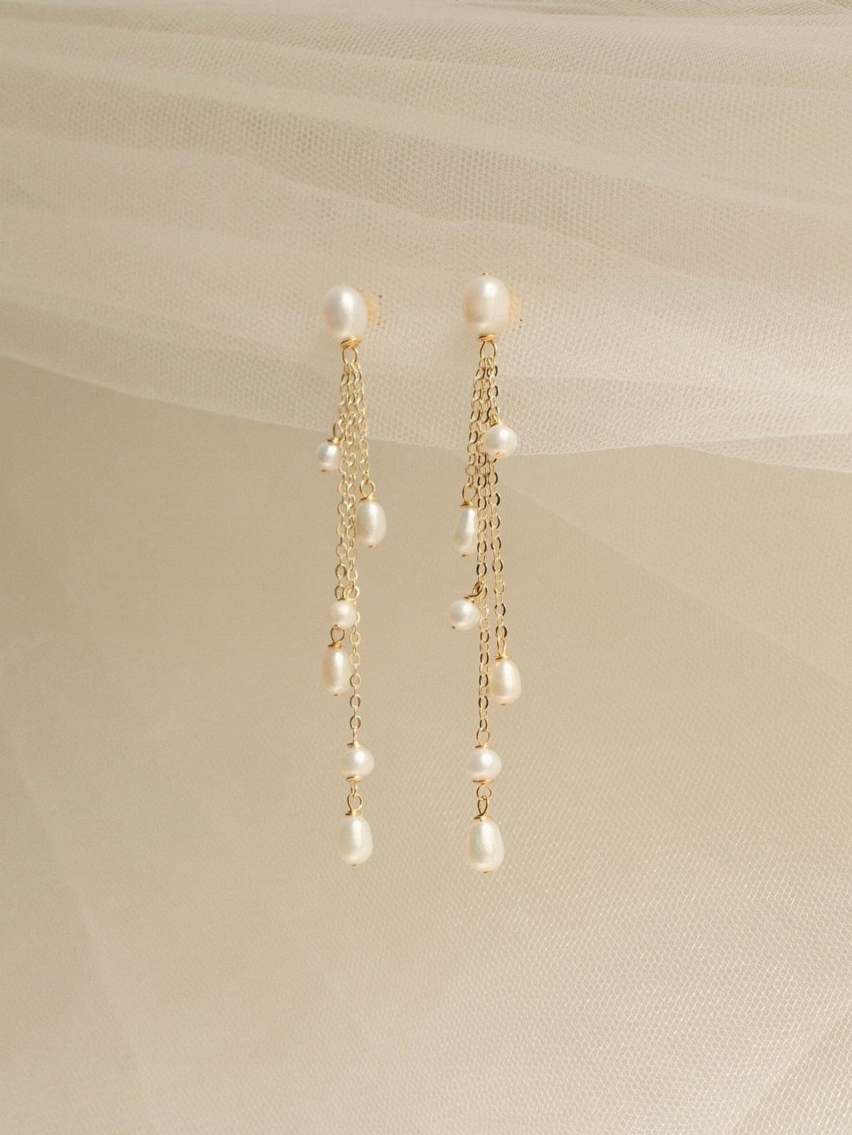 Long gold chain earrings with hanging white pearls against a cream fabric background.