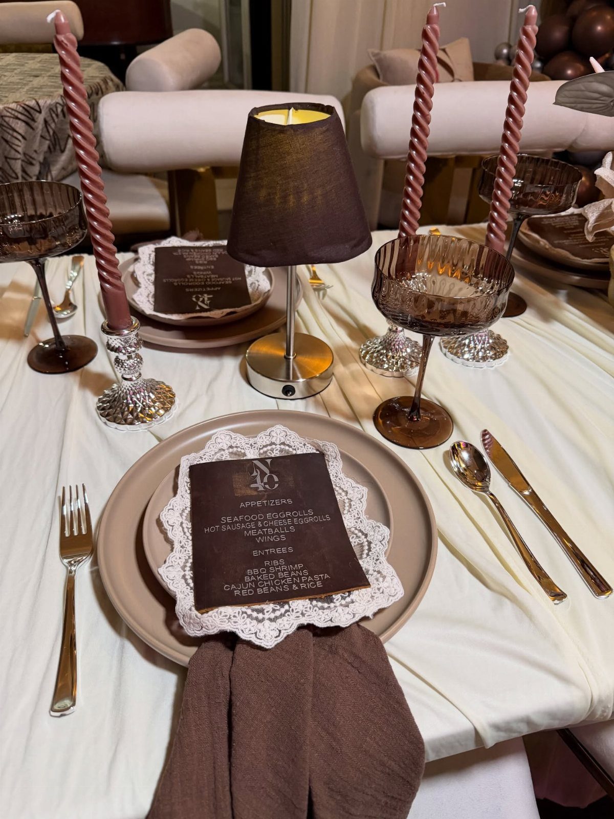 A formal table setting with tan plates, brown napkins, and tall pink candles on white linen.
