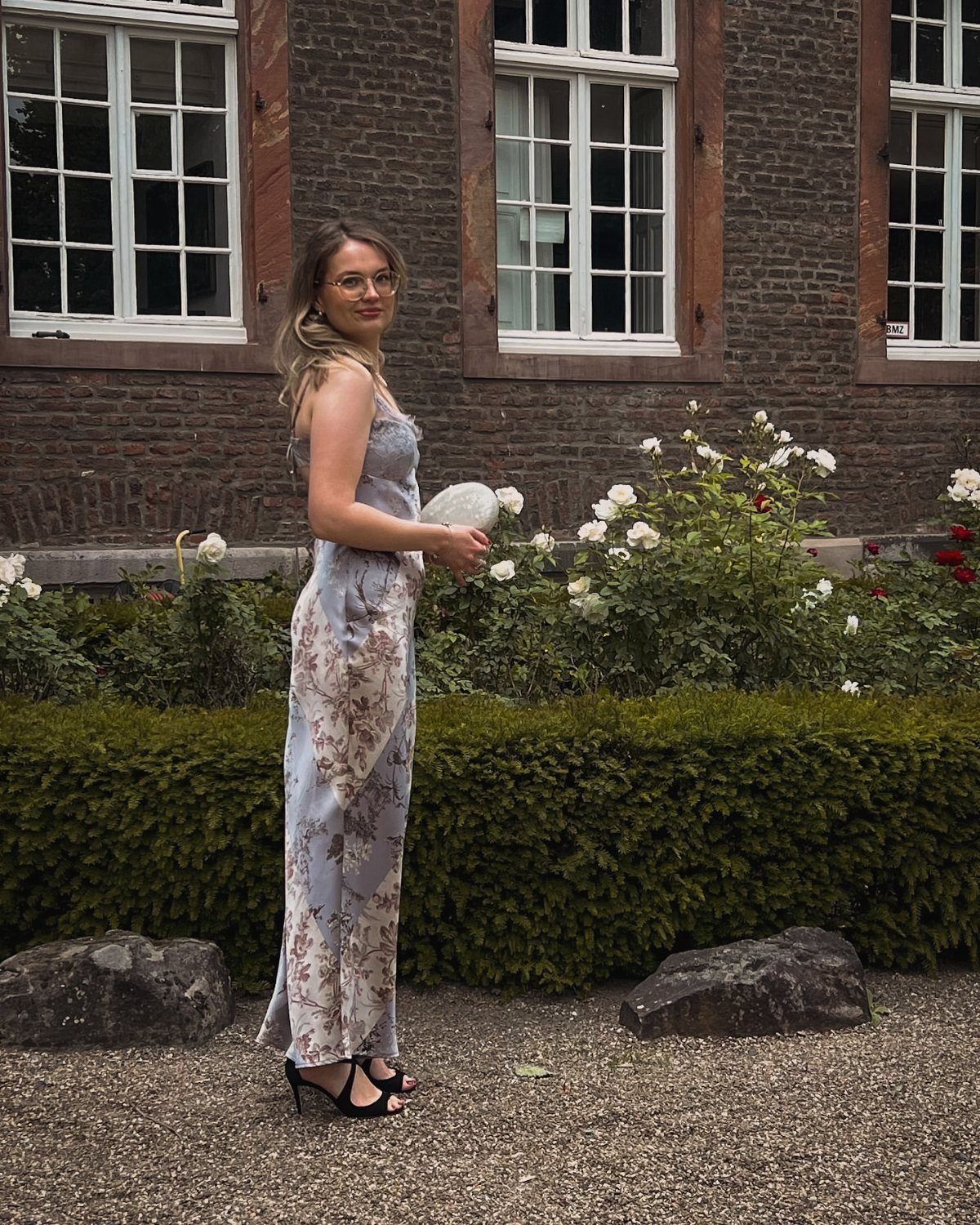 A woman wearing a light blue floral maxi dress and black sandals standing in a garden.
