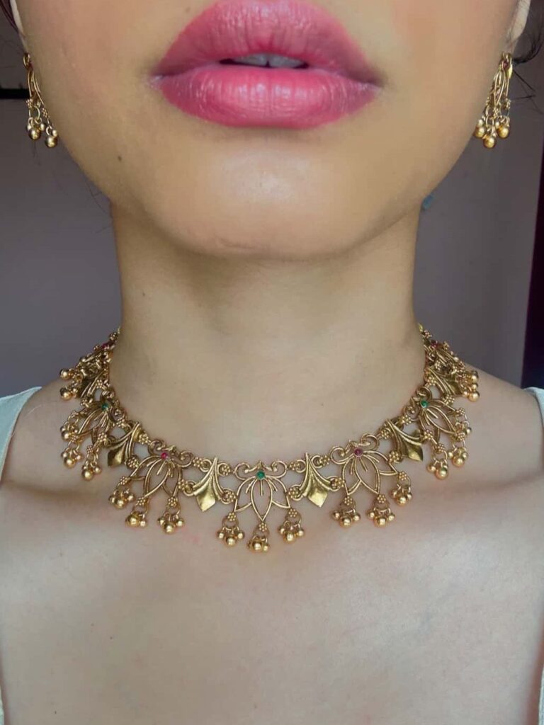 A person wearing a detailed gold choker necklace with small hanging bell charms and colorful stones.