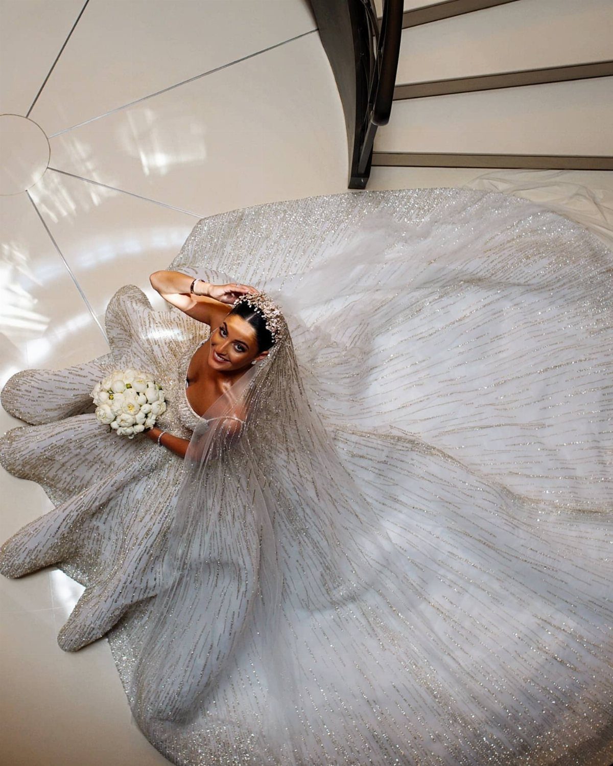 A bride in a shimmering silver gown poses on a staircase holding a white bouquet.