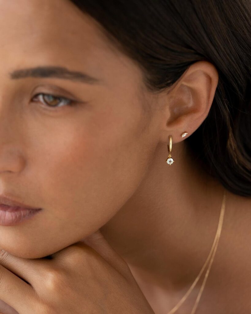 A woman wearing a small gold huggie earring with a dangling diamond charm.