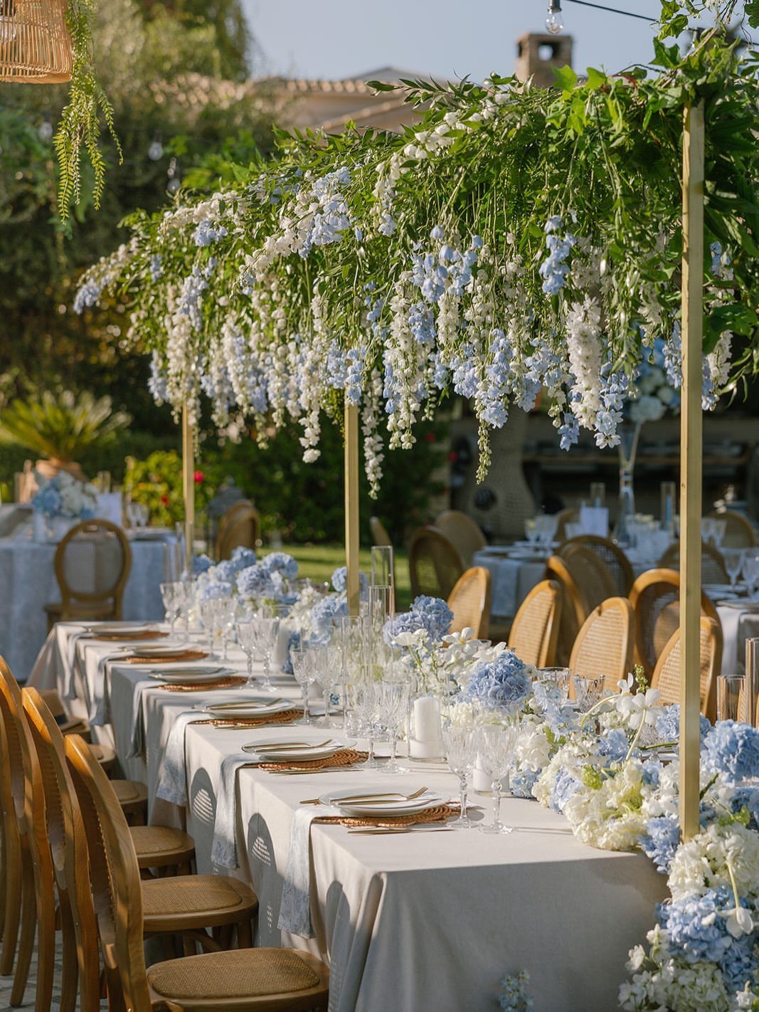 Long wedding reception table decorated with light blue flowers and white linens under a floral canopy.