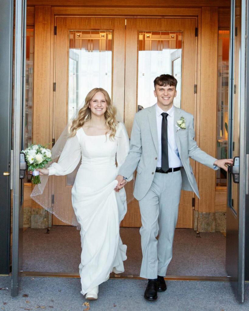 A bride in a white long-sleeved dress and a groom in a grey suit walking through doors.