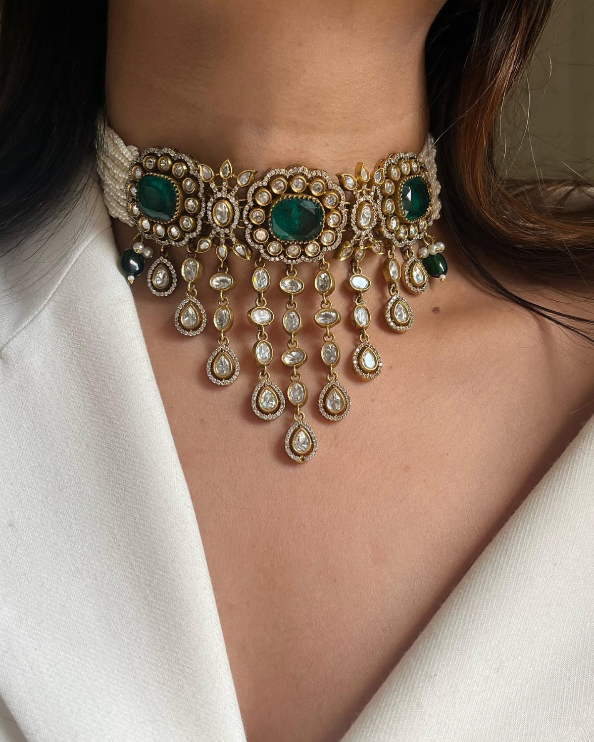 A woman wearing a gold choker necklace with large emeralds and dangling polki diamond stones.