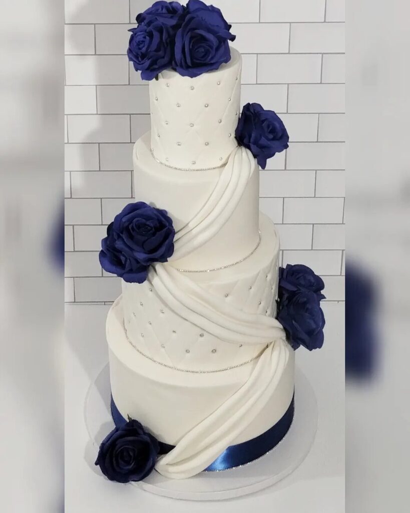 A four-tier white wedding cake decorated with royal blue roses and silver pearls.