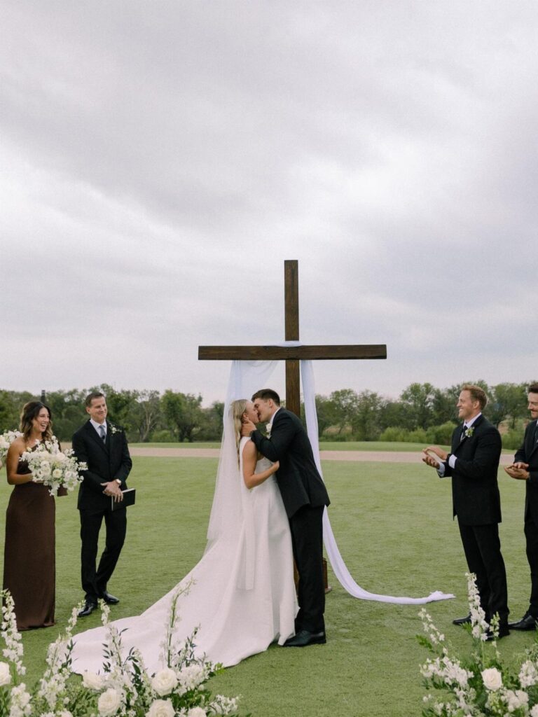A bride and groom kissing in front of a large wooden cross decorated with white fabric.