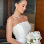 A smiling bride wearing a pearl necklace and earrings while holding a white flower bouquet.