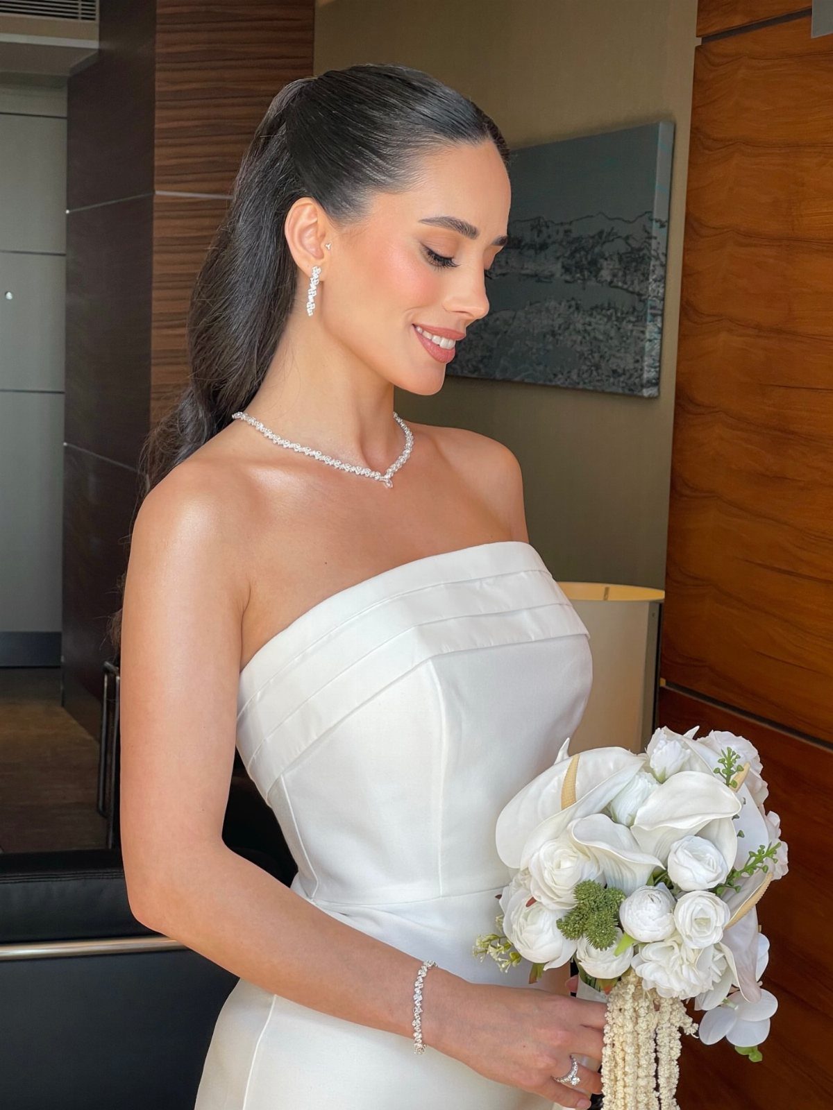 A smiling bride wearing a pearl necklace and earrings while holding a white flower bouquet.