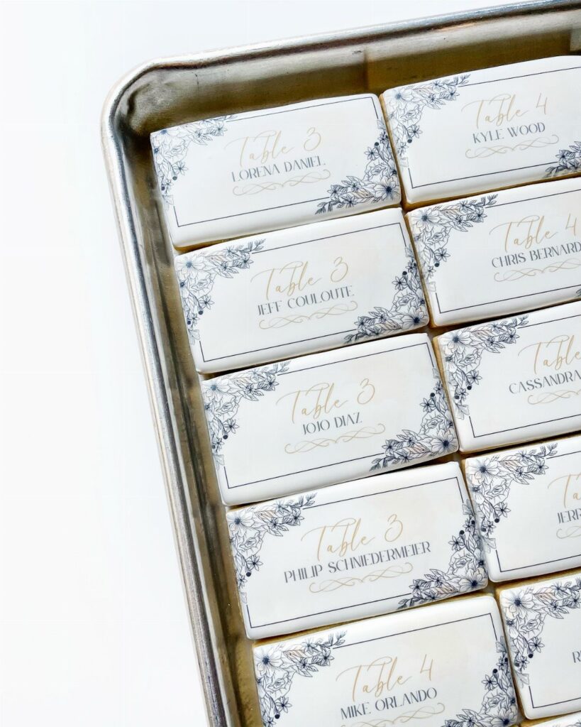 Rectangular white cookies with floral designs and gold calligraphy guest names on a silver tray.