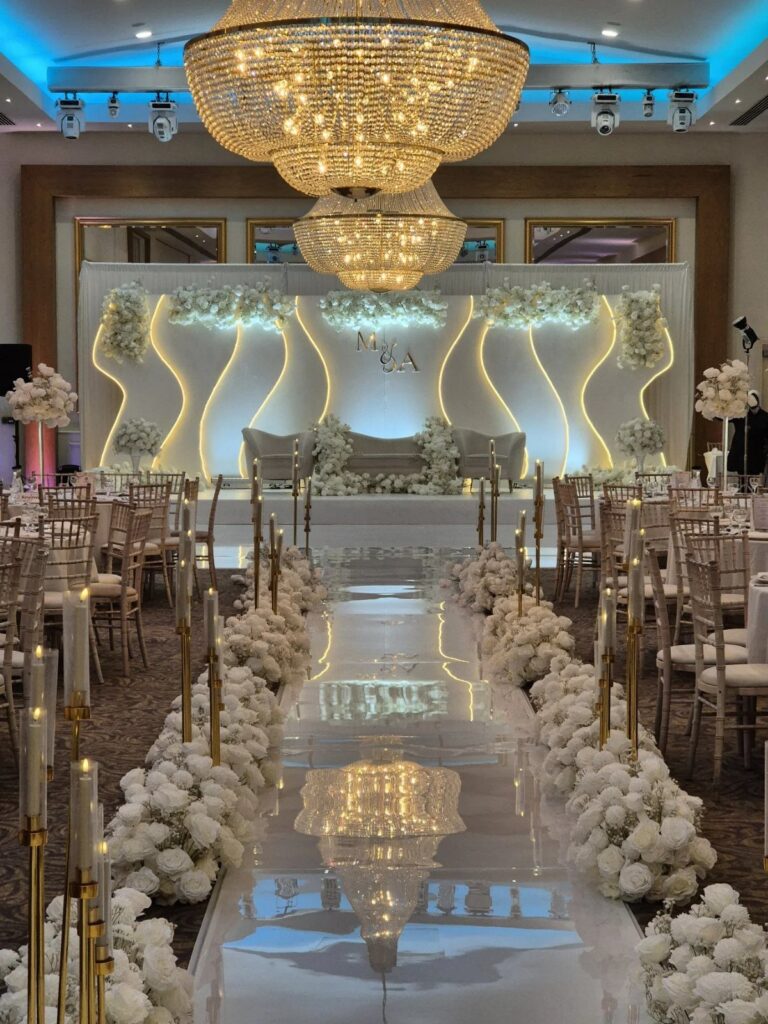White floral arrangements line a reflective wedding aisle with gold candle holders and large crystal chandeliers.