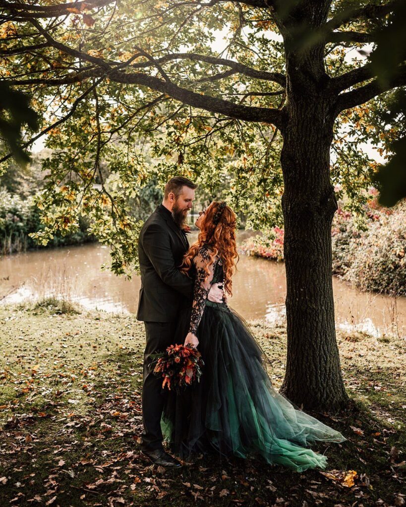 A bride in a dark green and black tulle wedding dress and a groom standing under a tree.