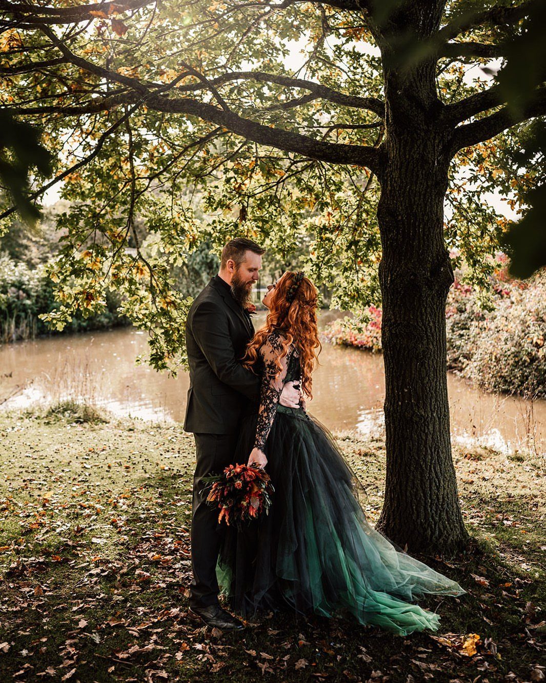A bride in a dark green and black tulle wedding dress and a groom standing under a tree.